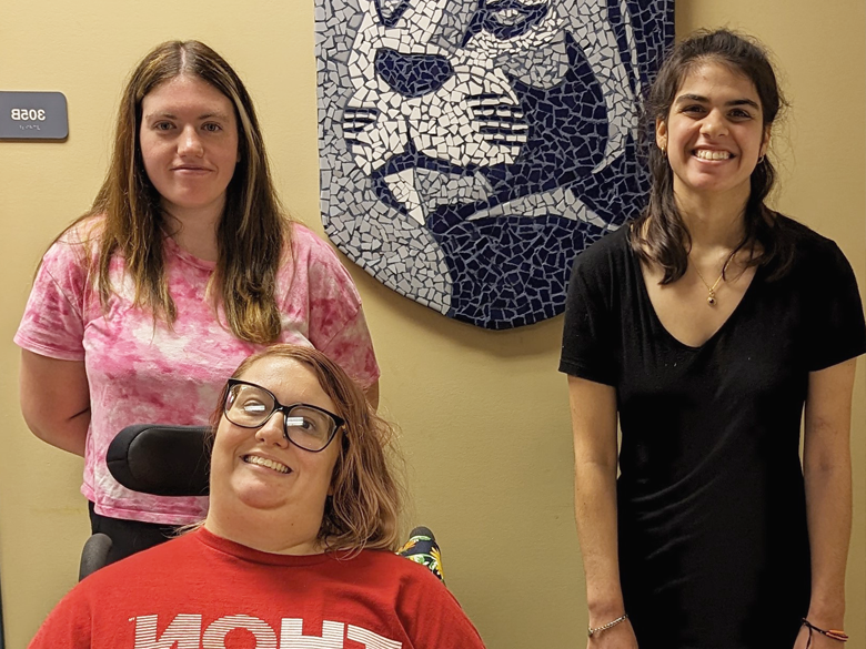 Three women standing in from of a Nittany Lion wall mosaic.